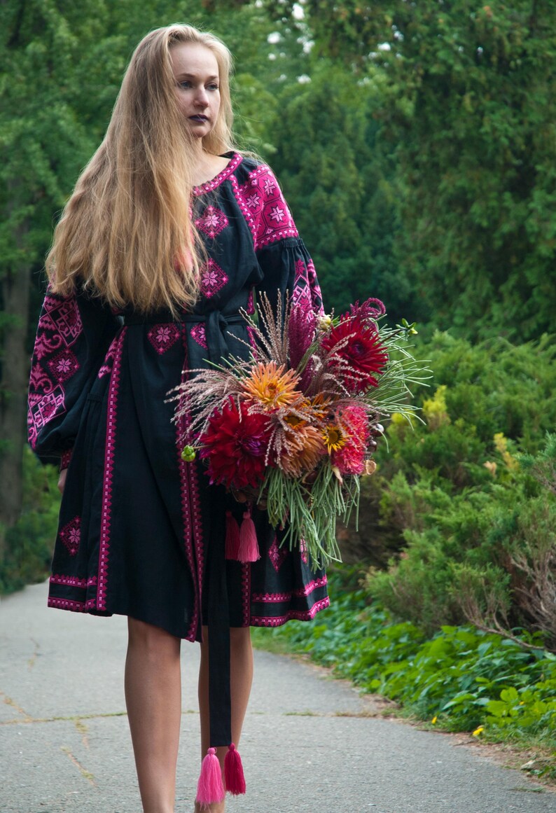 Op de afbeelding: Een zwarte jurk tot op de knie met lange mouwen en felroze geometrische borduurpatronen. Het kledingstuk heeft een bijpassende ceintuur met roze kwastjes en een ronde halslijn. Een groot, kleurrijk boeket dahlia's wordt aan de voorzijde gehouden.