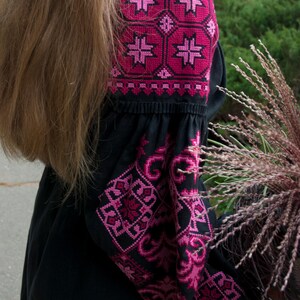 Op de afbeelding: Een zwarte blouse met lange mouwen en ingewikkelde geometrische borduursels in magenta en donkerroze op de schouders en mouwen. Het kledingstuk heeft een geplooide halslijn en wijde mouwen met gedetailleerde kruissteekpatronen in rozetinten.