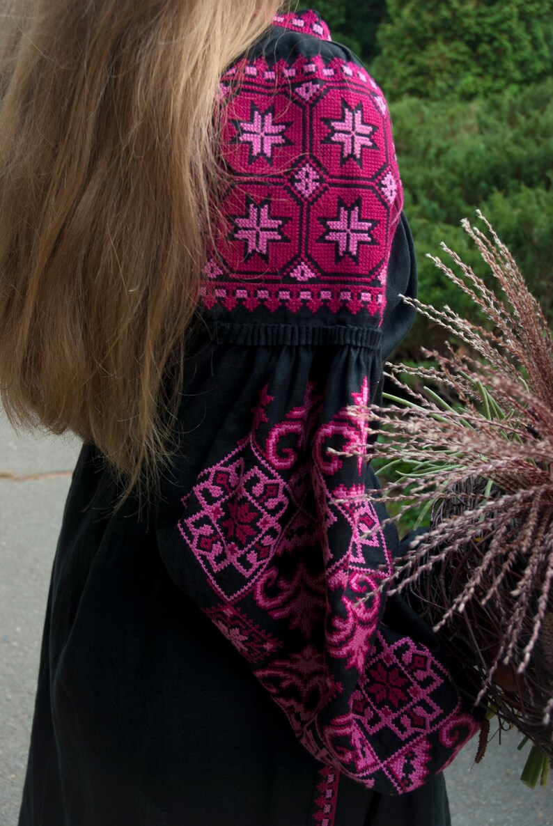 Op de afbeelding: Een zwarte blouse met lange mouwen en ingewikkelde geometrische borduursels in magenta en donkerroze op de schouders en mouwen. Het kledingstuk heeft een geplooide halslijn en wijde mouwen met gedetailleerde kruissteekpatronen in rozetinten.