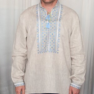 May include: A long-sleeved beige linen shirt featuring a vertical chest panel with blue and yellow geometric embroidery. The shirt has a mandarin collar with a drawstring closure finished with light blue tassels, and matching embroidered cuffs.