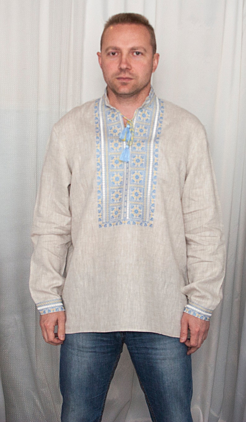 May include: A long-sleeved beige linen shirt featuring a vertical chest panel with blue and yellow geometric embroidery. The shirt has a mandarin collar with a drawstring closure finished with light blue tassels, and matching embroidered cuffs.