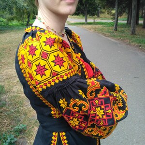Op de afbeelding: Een zwarte blouse met lange mouwen en ingewikkeld borduurwerk in levendige gele en rode geometrische patronen. Het kledingstuk heeft geplooide mouwen, een ronde halslijn met rode kwastjes en een bijpassende stoffen ceintuur in de taille.
