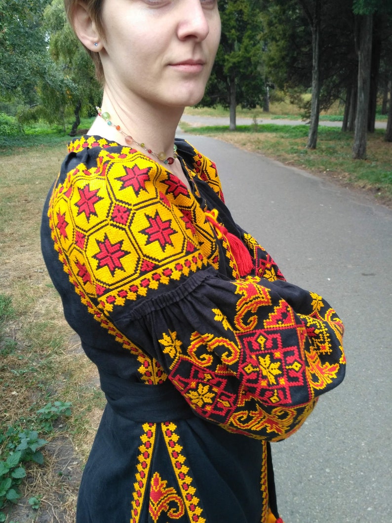 Op de afbeelding: Een zwarte blouse met lange mouwen en ingewikkeld borduurwerk in levendige gele en rode geometrische patronen. Het kledingstuk heeft geplooide mouwen, een ronde halslijn met rode kwastjes en een bijpassende stoffen ceintuur in de taille.
