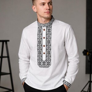 May include: White long-sleeve shirt with black and white embroidered detailing on the collar, placket, and cuffs. The shirt features a mandarin collar and wooden buttons. The design incorporates geometric and floral patterns.