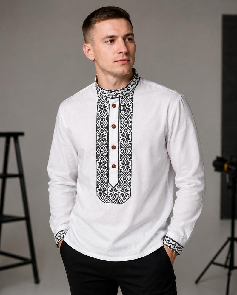 May include: White long-sleeve shirt with black and white embroidered detailing on the collar, placket, and cuffs. The shirt features a mandarin collar and wooden buttons. The design incorporates geometric and floral patterns.
