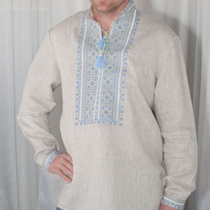 May include: A long-sleeved beige linen shirt featuring a vertical panel of intricate blue and yellow geometric embroidery down the center chest. The shirt includes a mandarin collar with a drawstring closure finished with two light blue tassels.