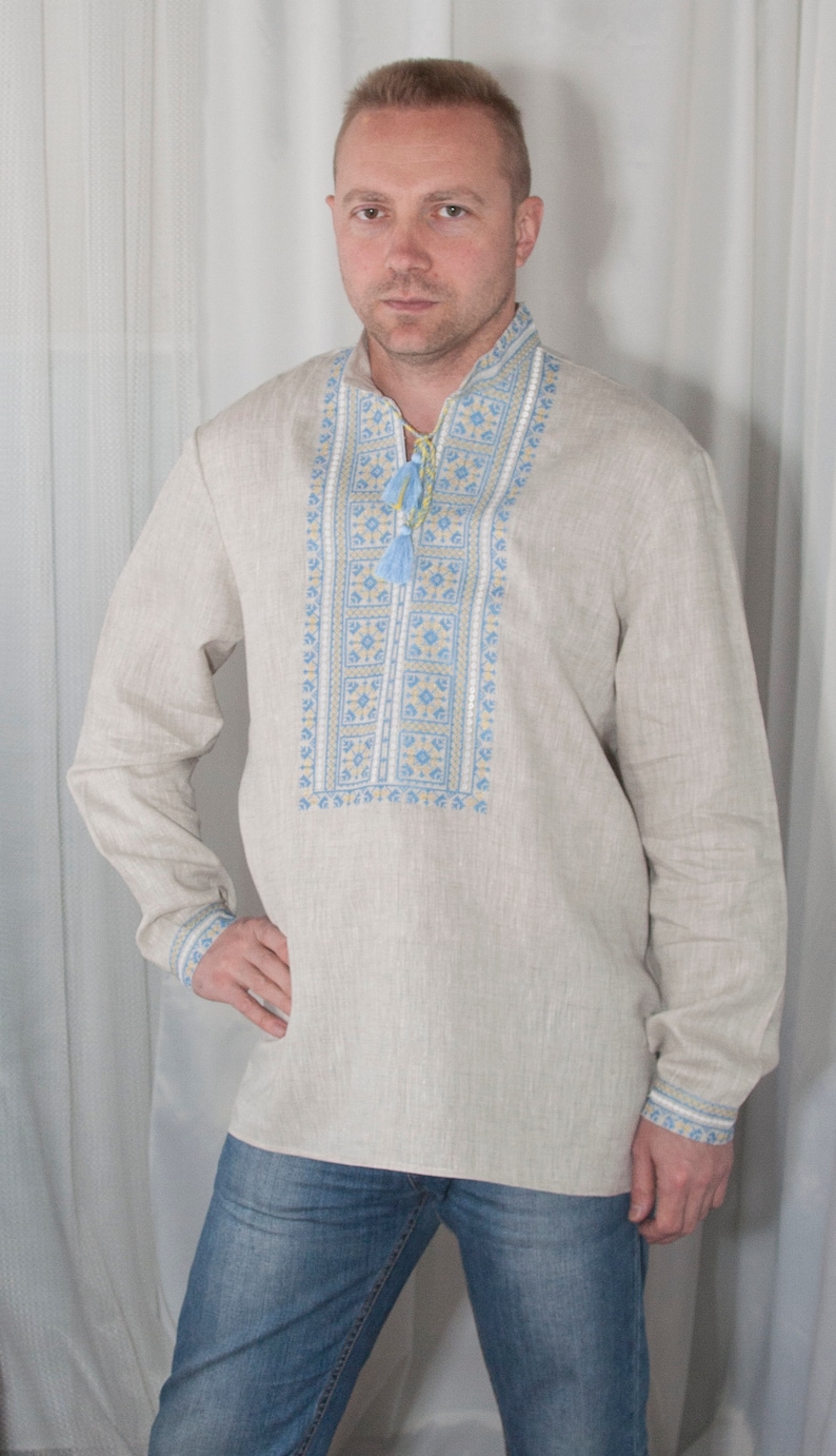 May include: A long-sleeved beige linen shirt featuring a vertical panel of intricate blue and yellow geometric embroidery down the center chest. The shirt includes a mandarin collar with a drawstring closure finished with two light blue tassels.