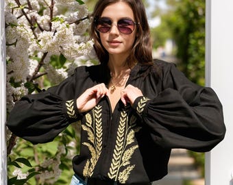 May include: A black long-sleeved blouse featuring gold-colored embroidery in a leaf and wheat pattern along the front placket and cuffs. The garment has a V-neckline, voluminous sleeves, and is paired with light blue denim jeans and sunglasses.