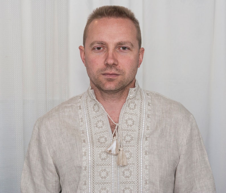 May include: A beige linen shirt featuring a stand-up collar with a V-neck opening and decorative white embroidery along the front placket. The shirt includes a drawstring closure at the neckline finished with two small, matching beige tassels.