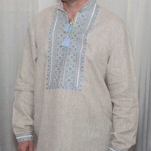 May include: A beige long-sleeved linen shirt featuring a stand-up collar and a central vertical panel with intricate blue and yellow geometric embroidery. The neckline includes a drawstring with light blue tassels, and the cuffs feature matching embroidery.