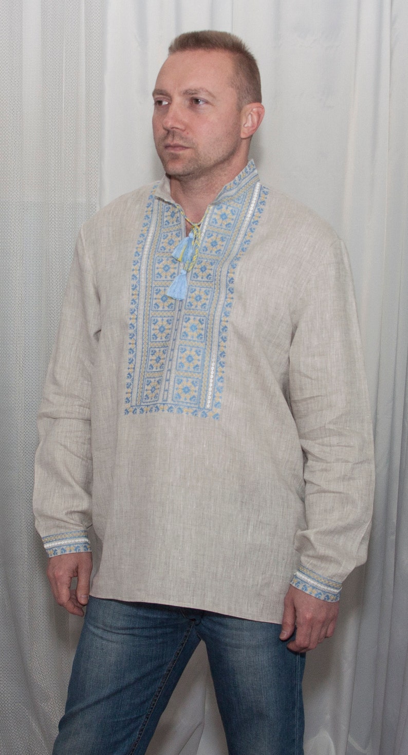 May include: A beige long-sleeved linen shirt featuring a stand-up collar and a central vertical panel with intricate blue and yellow geometric embroidery. The neckline includes a drawstring with light blue tassels, and the cuffs feature matching embroidery.