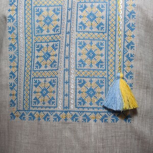May include: A light gray linen shirt featuring intricate cross-stitch embroidery in bright blue and yellow thread. The geometric pattern includes square motifs and vertical borders, finished with a braided cord and a two-toned blue and yellow tassel.