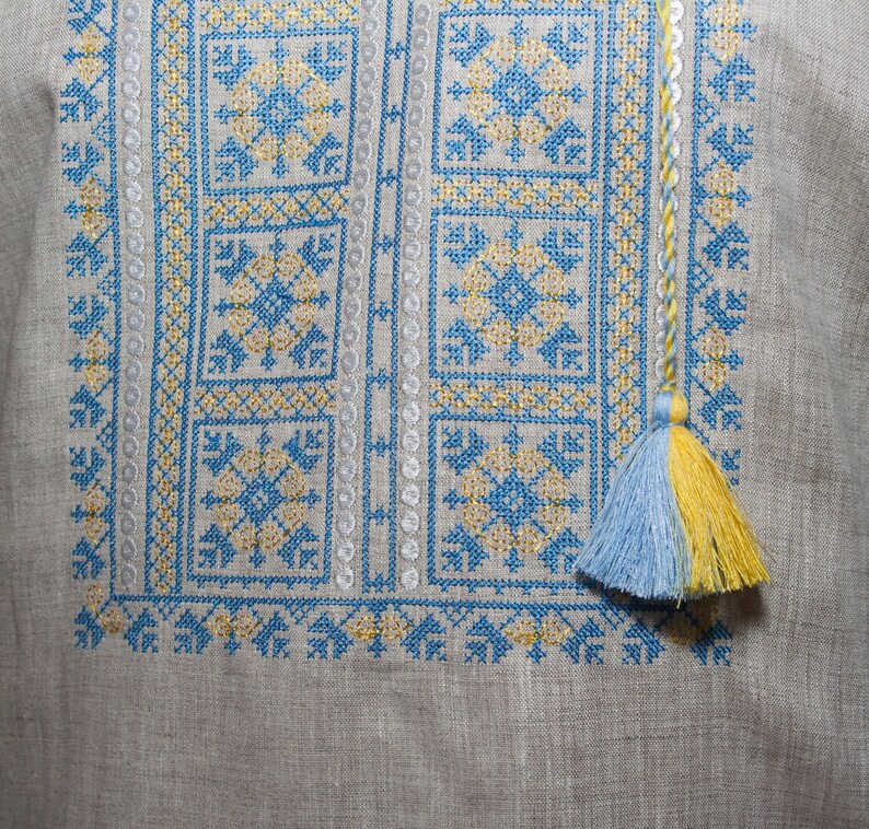 May include: A light gray linen shirt featuring intricate cross-stitch embroidery in bright blue and yellow thread. The geometric pattern includes square motifs and vertical borders, finished with a braided cord and a two-toned blue and yellow tassel.