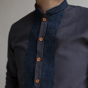 May include: Charcoal gray shirt with a mandarin collar and a decorative navy blue embroidered panel down the front. The shirt features wooden buttons and a detailed geometric pattern on the collar and panel.