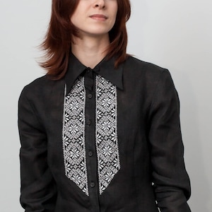 Puede incluir: Una camisa negra con cuello puntiagudo y mangas largas. La camisa presenta un diseño bordado en blanco y negro en la parte delantera. La mujer de la foto tiene el pelo rojo.
