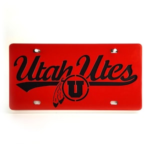 May include: A red license plate with the words "Utah Utes" in black script. The plate features a black circle with a "U" inside and a feather graphic. The plate has four holes for mounting.