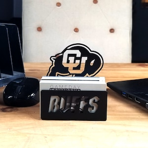 May include: Black metal business card holder with a cutout of the University of Colorado Buffaloes logo and the text "GAMEDAY IRONWORKS BUFFS EST. 2014".