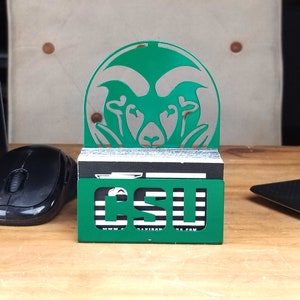 May include: Green metal business card holder with a cutout of a ram's head and the text "CSU" on the front. The holder is sitting on a wooden surface.