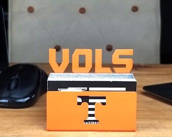 May include: Orange and black business card holder with the word "VOLS" in large orange letters and a stylized orange "T" with black and white stripes. The holder has the word "DAYIRON" printed on the bottom.