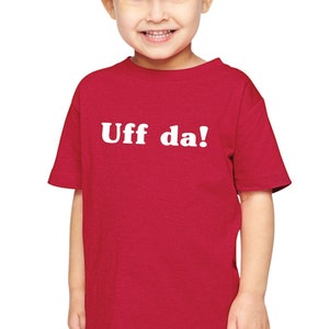 May include: A young child wearing a red t-shirt with the white text "Uff da!" in a playful font. The child is wearing blue jeans and has their hair up in a bun. The background is plain white.
