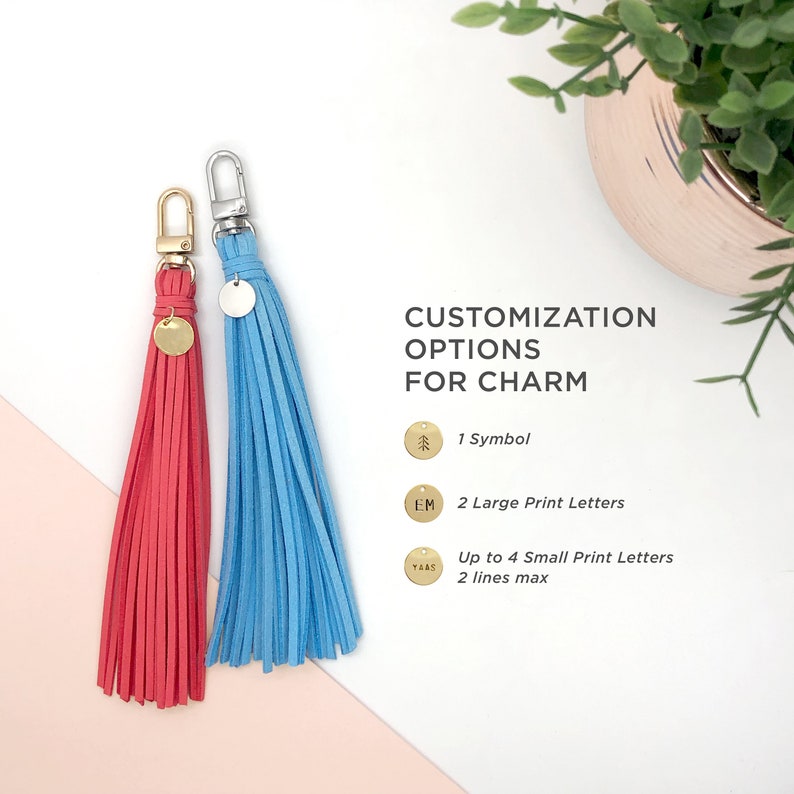 May include: Two tassel keychains with different colored tassels, one red and one blue. The keychains have a small round charm with different customization options: a pine tree, the letters "EM", and the word "YAAS". The text "CUSTOMIZATION OPTIONS FOR CHARM" is written above the charms. The text below the charms describes the customization options: 1 Symbol, 2 Large Print Letters, Up to 4 Small Print Letters, 2 lines max.