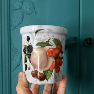 Peut inclure: Pot en céramique blanche avec un motif floral représentant des cerises, des pêches et des mûres. Le pot a une forme arrondie et une large ouverture.