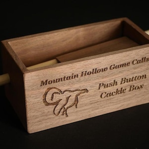 May include: A wooden push button cackle box with the text "Mountain Hollow Game Calls" and a ram graphic. The box is rectangular with a sliding lid and a wooden dowel on each side. The wood has a natural grain.