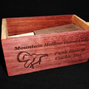 May include: A wooden box with a red-stained finish. The box is engraved with the text "Mountain Hollow Game Calls" and "Push Button Cackle Box". The box has a small wooden stick protruding from one side.