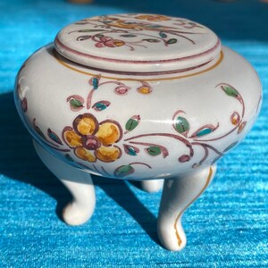 May include: A white ceramic trinket box with a floral design in shades of yellow, pink, green, and brown. The box has three legs and a lid.