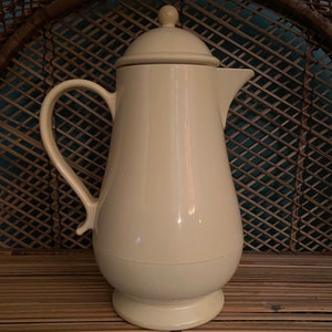 May include: A cream-colored ceramic pitcher with a lid and a handle. The pitcher is sitting on a woven surface.