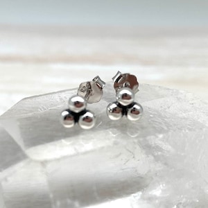 May include: A pair of small, silver stud earrings. Each earring has three small, round, polished silver beads in a triangular cluster. The earrings are displayed on a light-coloured, textured surface.
