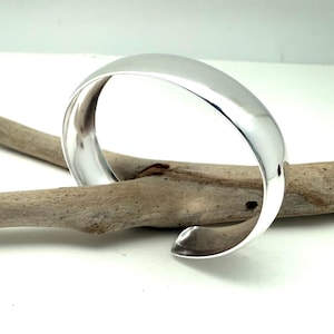 May include: A polished silver cuff bracelet with a smooth, curved design. The bracelet is resting on a piece of driftwood, showcasing its reflective surface. The open design allows for easy wear. A simple, elegant piece of jewelry.