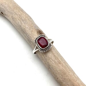 Op de afbeelding: Een zilveren ring met een donkerrode edelsteen gezet in een geperlde bezel. De ring ligt op een stuk drijfhout.