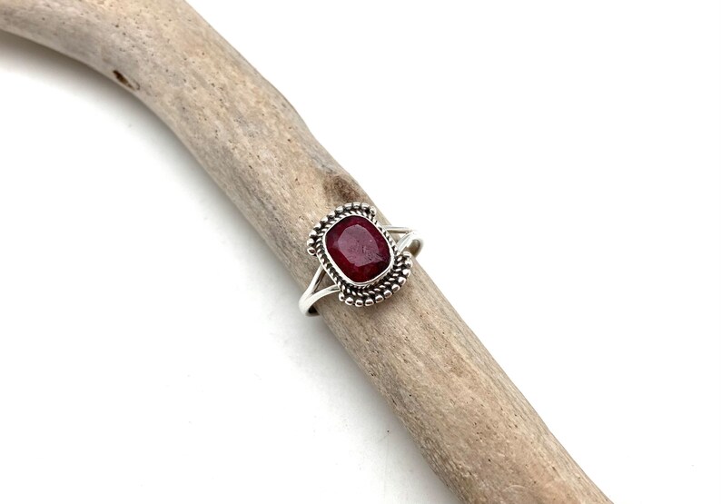 Op de afbeelding: Een zilveren ring met een donkerrode edelsteen gezet in een geperlde bezel. De ring ligt op een stuk drijfhout.