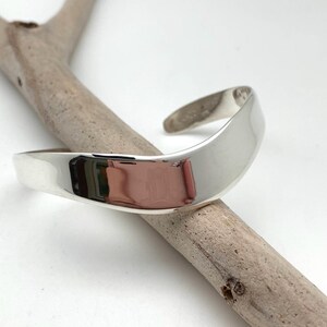 May include: A silver bangle bracelet with a curved, smooth design. The bracelet is made of polished metal and has a simple, elegant look.