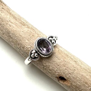 May include: A silver ring with an oval-shaped amethyst gemstone set in a beaded bezel. The ring has a delicate design with small silver beads on the band.