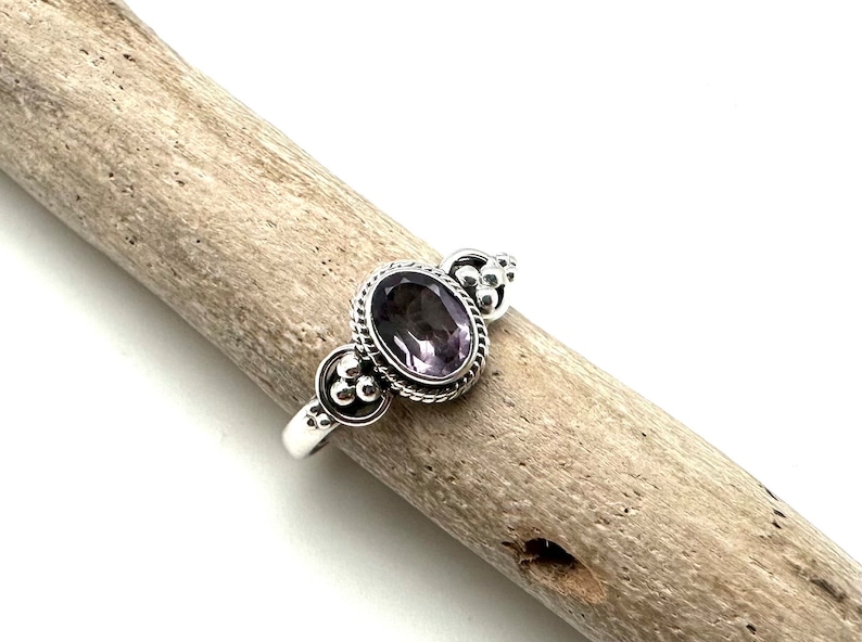 May include: A silver ring with an oval-shaped amethyst gemstone set in a beaded bezel. The ring has a delicate design with small silver beads on the band.