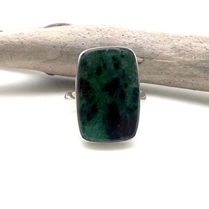 May include: A rectangular ring with a dark green stone featuring black inclusions, set in a silver band. The ring's stone has a polished surface and a rectangular shape. The ring is displayed on a white surface.