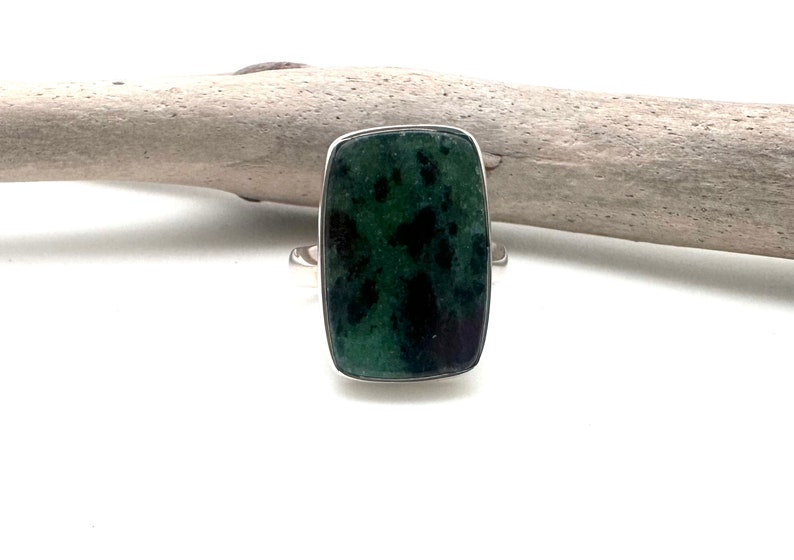 May include: A rectangular ring with a dark green stone featuring black inclusions, set in a silver band. The ring's stone has a polished surface and a rectangular shape. The ring is displayed on a white surface.