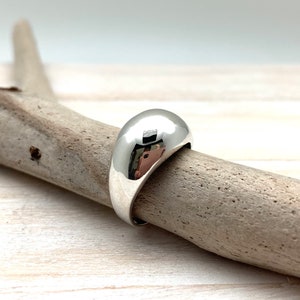 May include: A silver ring with a smooth, rounded dome shape. The ring is sitting on a piece of driftwood.