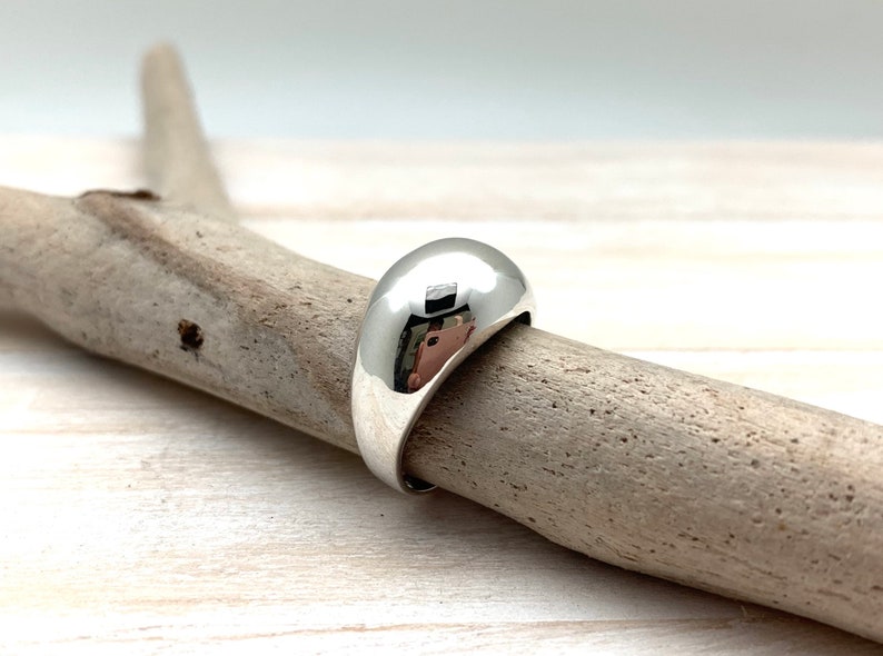 May include: A silver ring with a smooth, rounded dome shape. The ring is sitting on a piece of driftwood.