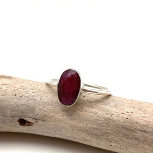 May include: A silver ring with a large, oval-shaped ruby gemstone. The ring is set on a piece of driftwood.