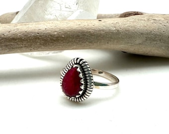 Puede incluir: Un anillo de plata con una piedra roja en forma de lágrima engastada en un engaste de plata detallado. El anillo tiene una banda texturizada.