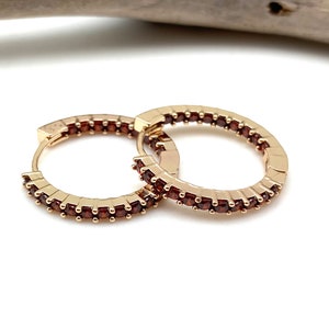 May include: Gold hoop earrings with a row of small, dark red gemstones around the edge.