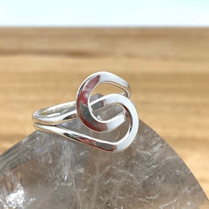 Puede incluir: Un anillo de plata con un diseño de nudo único. El anillo está sobre una superficie de cristal transparente.