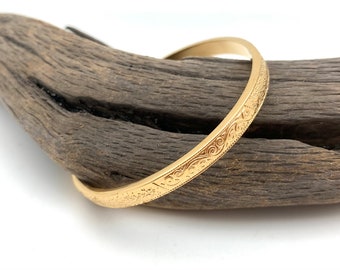 Puede incluir: Un brazalete de oro con un diseño intrincado grabado. El brazalete está sobre un trozo de madera a la deriva.