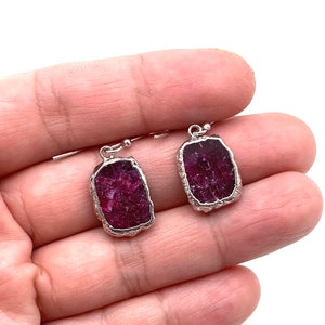 May include: A pair of silver earrings with raw ruby stones. The stones are a deep red color and have a rough, natural texture.