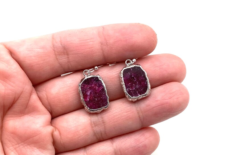 May include: A pair of silver earrings with raw ruby stones. The stones are a deep red color and have a rough, natural texture.