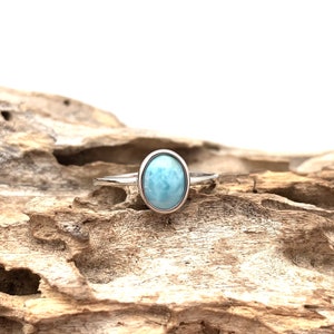 May include: A silver ring featuring a light blue oval-shaped gemstone. The ring is resting on a piece of driftwood.