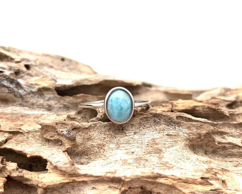 May include: A silver ring featuring a light blue oval-shaped gemstone. The ring is resting on a piece of driftwood.